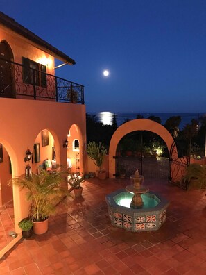 Un patio con una fuente, plantas en macetas y vista al mar al atardecer.