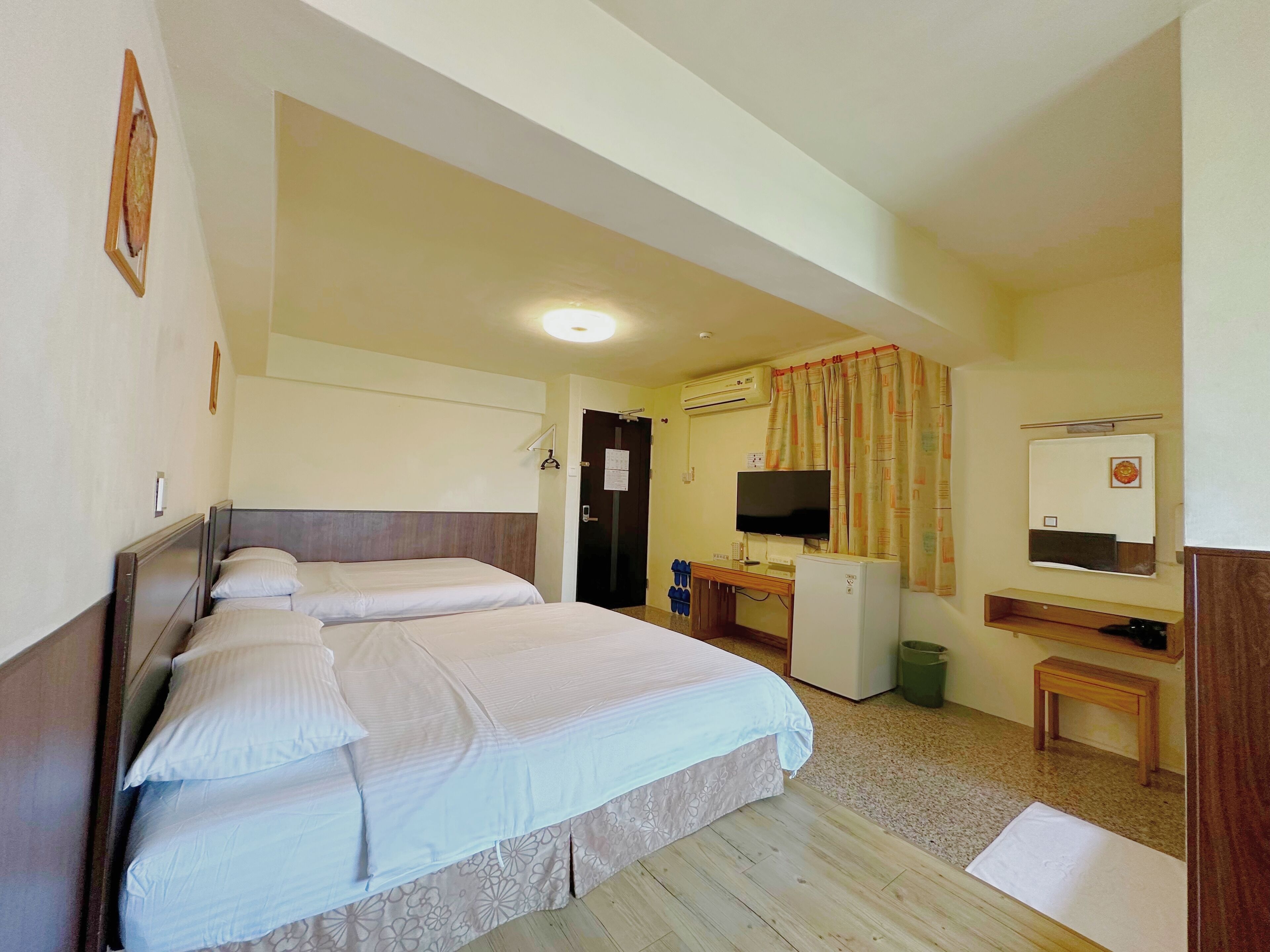 A hotel room with two beds, a desk with a computer, a television, and an air conditioning unit.