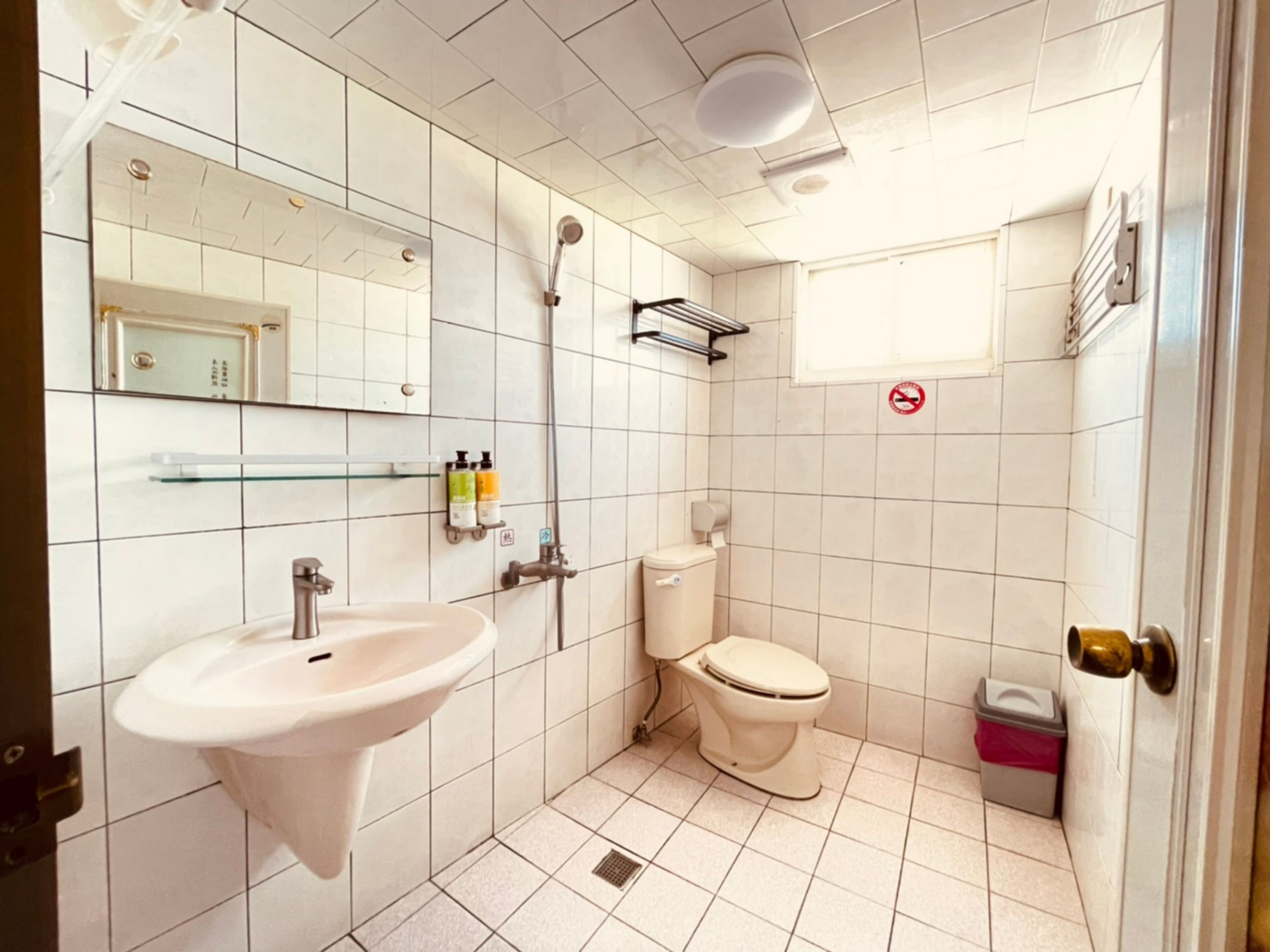 A small, clean bathroom with a sink, toilet, and shower. There is a mirror, a shelf with toiletries, and a trash bin.