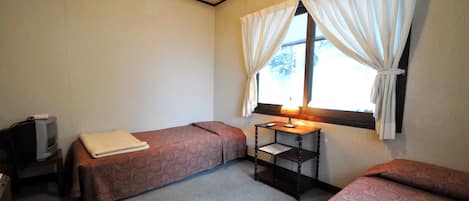 Una habitación pequeña y sencilla con dos camas, un escritorio con lámpara y una ventana con cortinas.