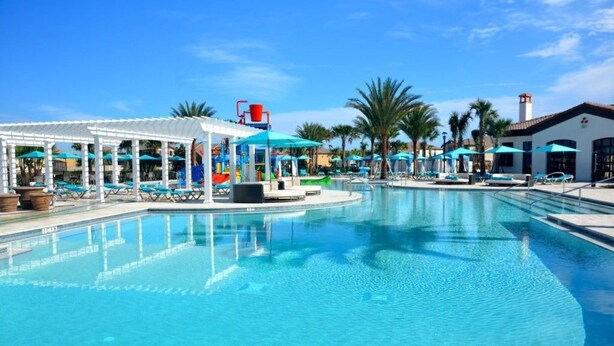 A swimming pool with lounge chairs and umbrellas, surrounded by palm trees and a covered area with a water slide.