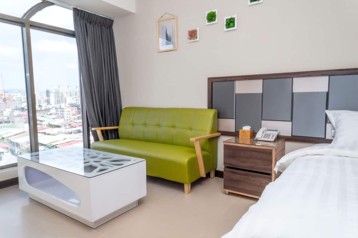 A modern hotel room with a green sofa, a glass coffee table, a wooden nightstand with a phone, and a bed with white bedding.