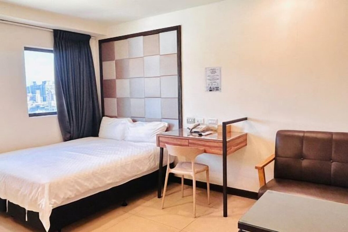 A hotel room with a bed, a desk with a chair, a sofa, and a city view through the window.