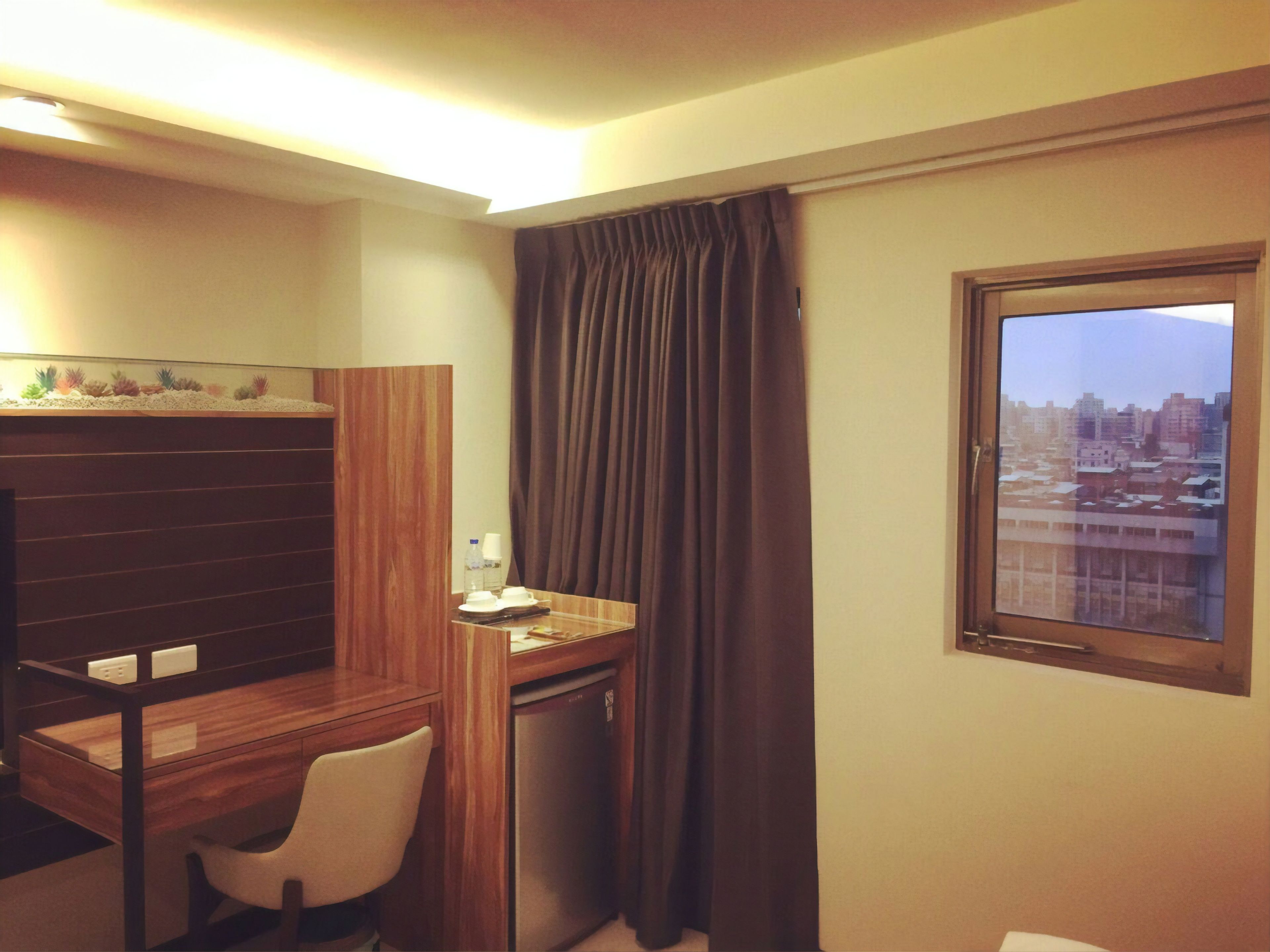 A compact hotel room with a wooden desk, a chair, a small refrigerator, and a window with a cityscape view.