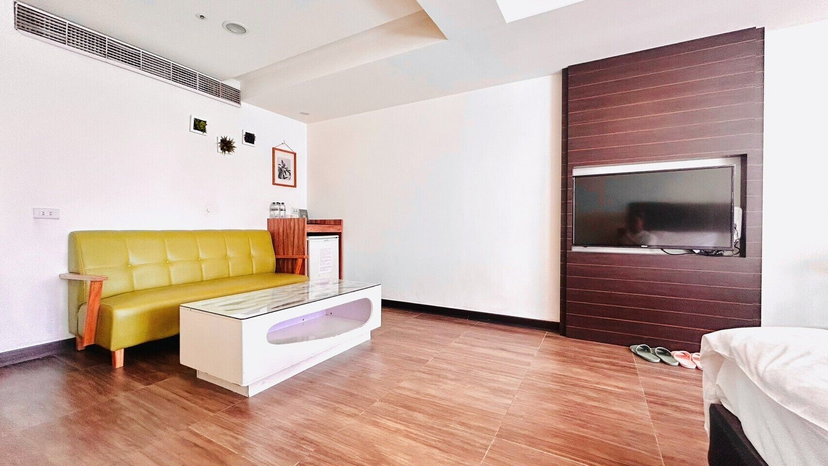 Una habitación de hotel moderna con un sofá amarillo, una mesa de centro de vidrio, un televisor de pantalla plana montado en un panel de madera y una cama con una colcha blanca.