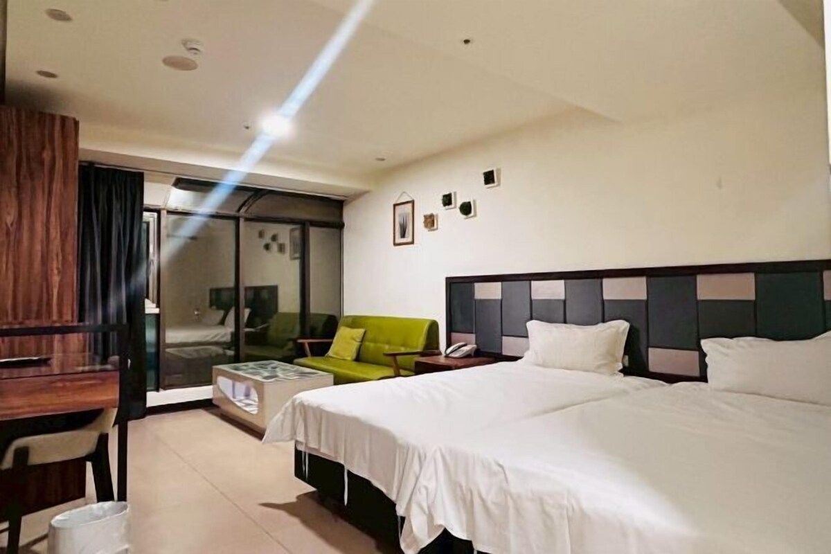 A hotel room with a large bed, a desk, a green armchair, and a sliding glass door leading to a balcony.
