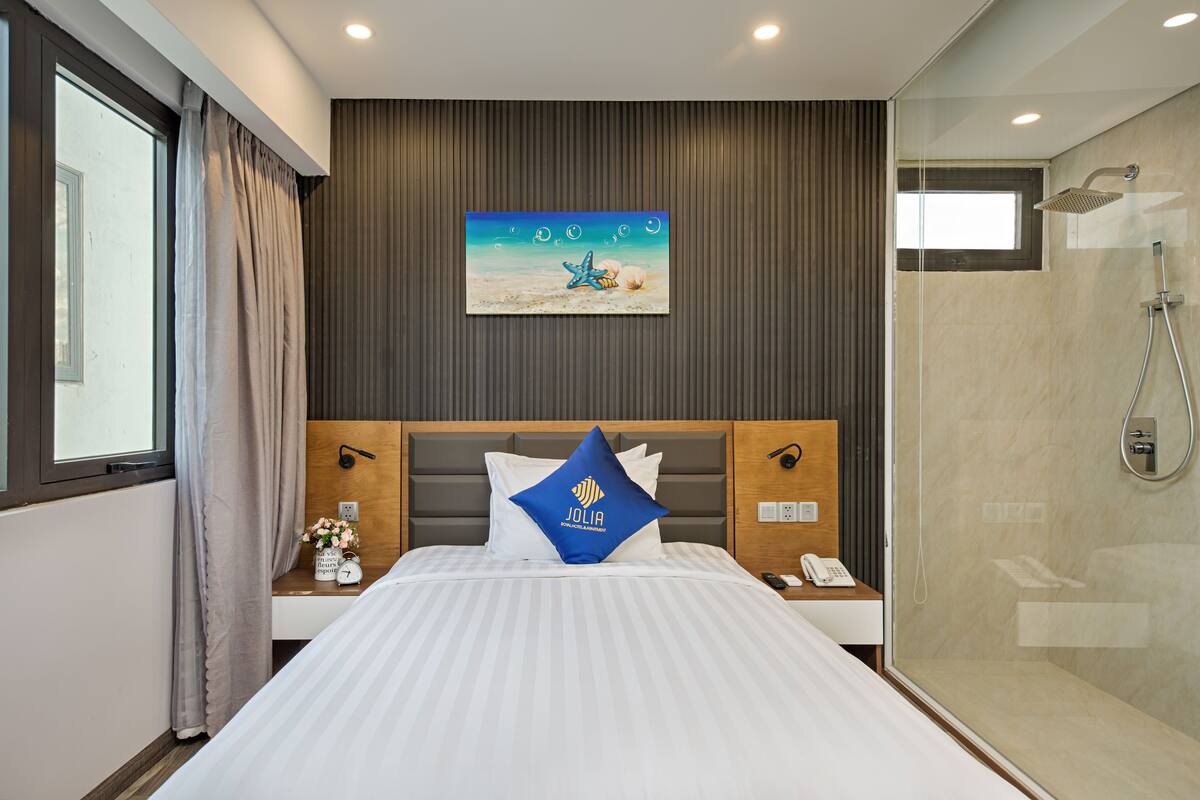 Habitación de hotel con cama, un cuadro de una escena de playa, ducha y mesita de noche con un teléfono y un florero.