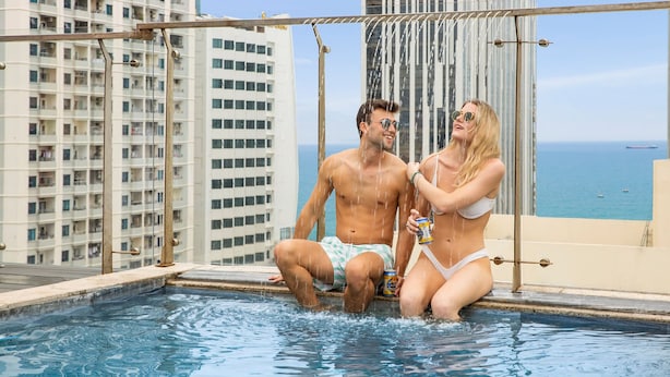 Una pareja en una piscina en la azotea con vista a la ciudad, una barandilla metálica y cielo despejado.