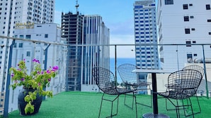 Una terraza en la azotea con césped artificial, dos sillas de malla metálica negra y una maceta con flores moradas.