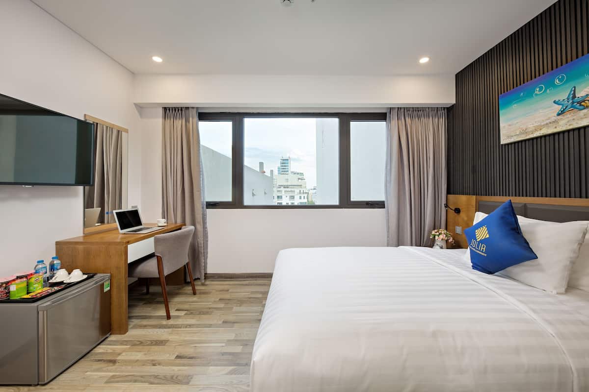 Habitación de hotel con cama, escritorio, ordenador portátil y vistas a edificios a través de la ventana.