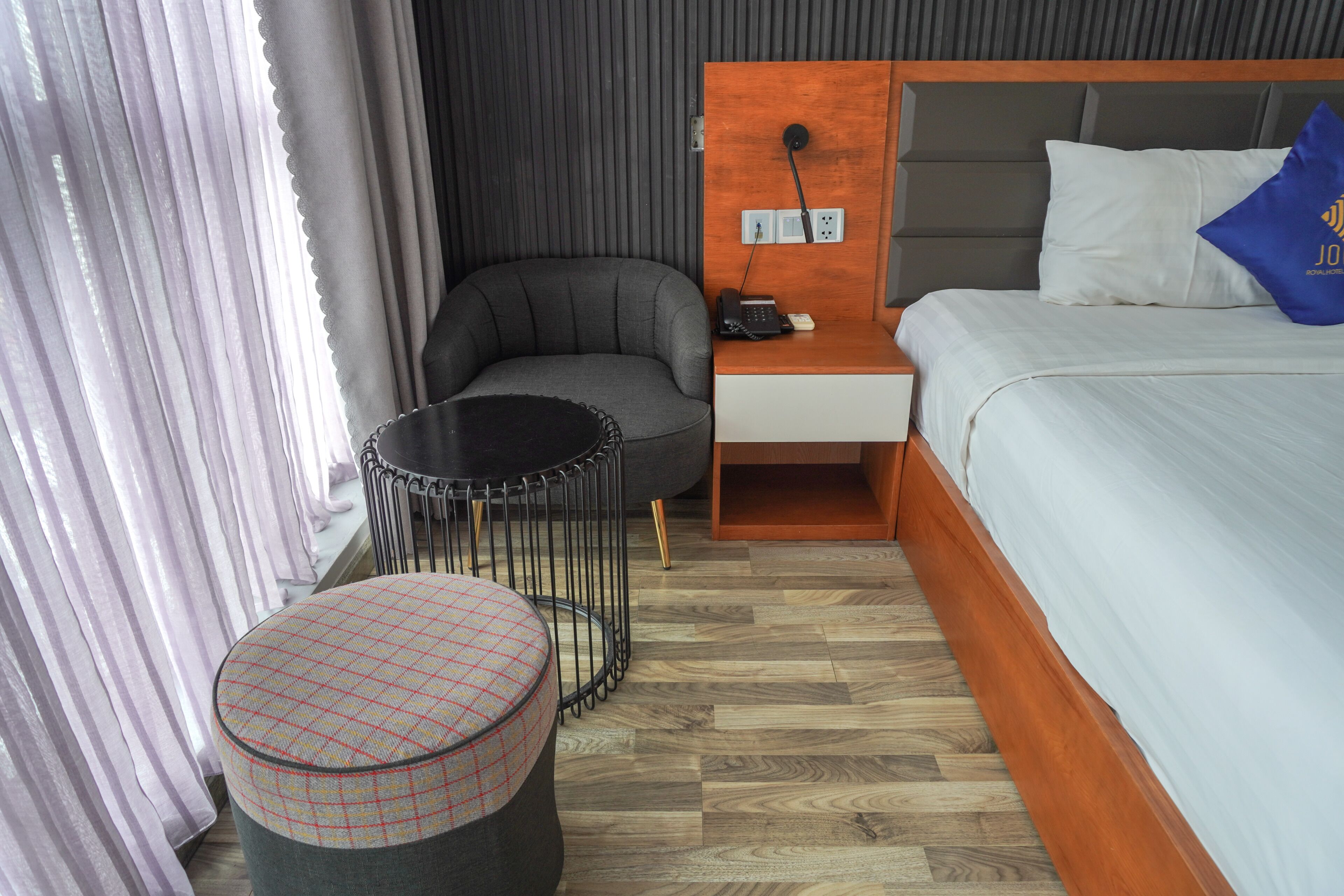 A hotel room with a bed, a desk, a chair, a round ottoman, and a curtain.