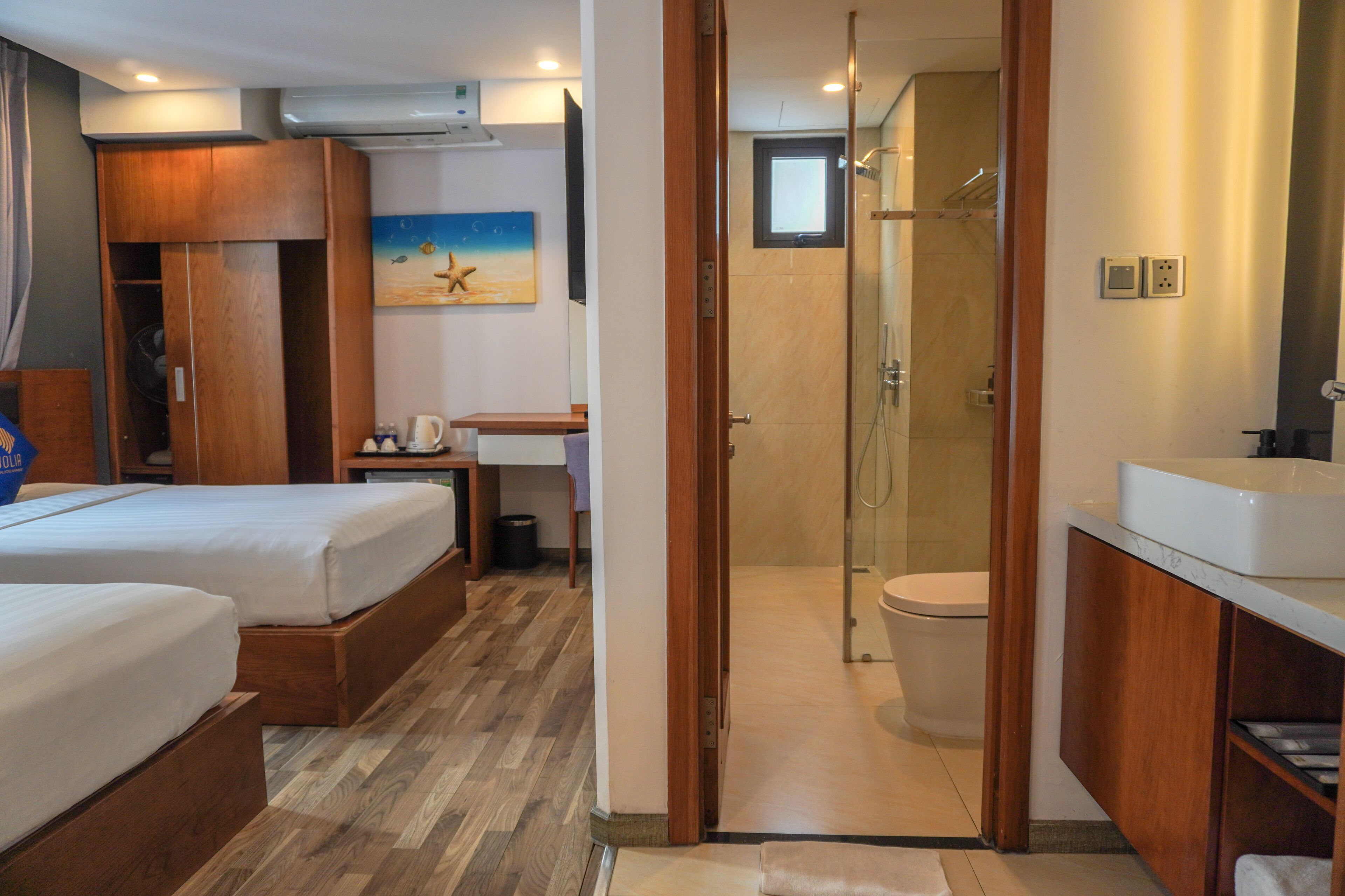 A hotel room with two beds, a wooden wardrobe, a painting, a bathroom with a shower, and a sink.