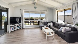 A spacious living room with a large TV, a sofa, and a coffee table. There is a ceiling fan, a bookshelf, and a view of the outdoors through large windows. - Sunset View- Magnificiant Ocean Views (Lancelin)