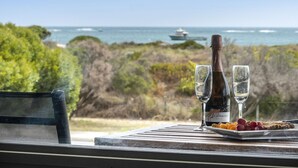 A table with a bottle of wine, two glasses, and a plate of fruit by a window overlooking the ocean. - Sunset View- Magnificiant Ocean Views (Lancelin)