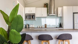 A modern kitchen with white cabinets, a stainless steel microwave, a kettle, and a toaster. There are three stools with dark cushions at the counter. - Sunset View- Magnificiant Ocean Views (Lancelin)