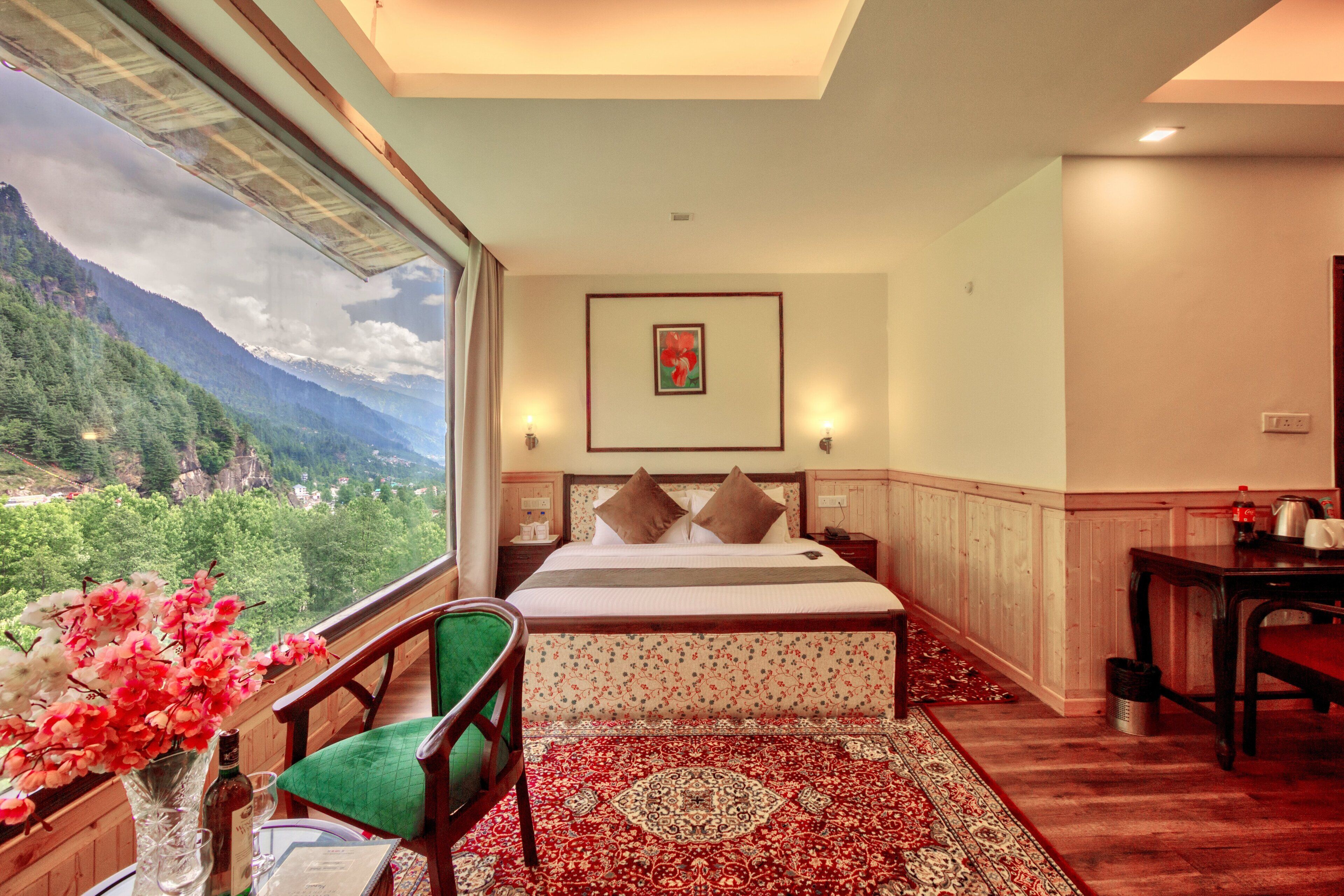 A hotel room with a large window offering a mountain view, a bed with a floral-patterned bedspread, a green chair, and a wooden table with a kettle.