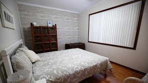 Une chambre à coucher avec un lit, une armoire en bois, une fenêtre avec des stores, une petite table et une lampe.