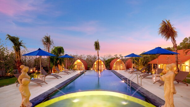 Zona de piscina con sillas de descanso y sombrillas, rodeada de palmeras y un cielo al atardecer.