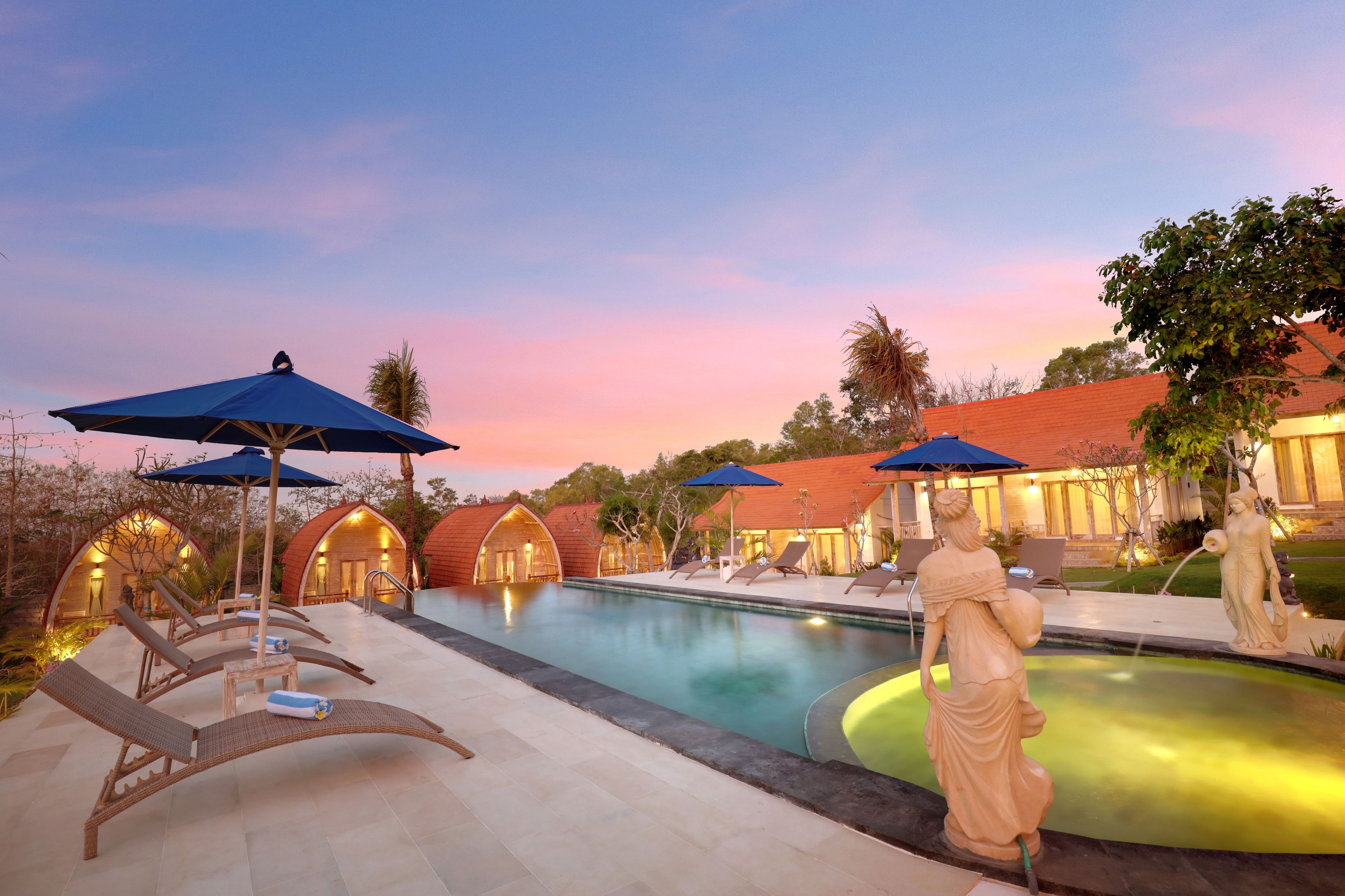 Un área de piscina con sillones de descanso y sombrillas, rodeada de edificios con techos abovedados.