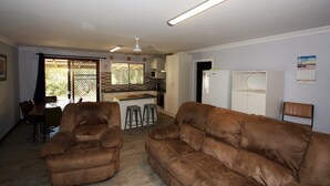 A living room with two brown sofas, a dining table with chairs, a kitchen area with a refrigerator and a microwave, and a ceiling fan.