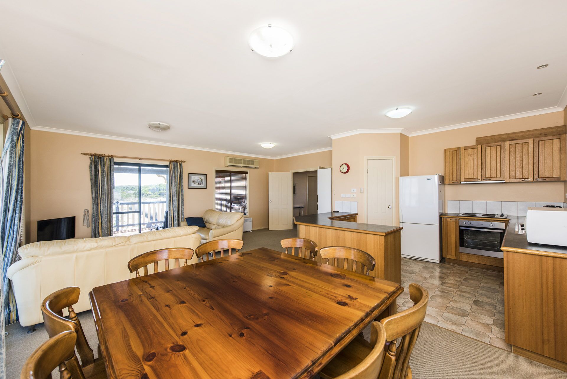 Un'ampia zona giorno con tavolo da pranzo in legno, divano beige e cucina con mobili e elettrodomestici in legno.