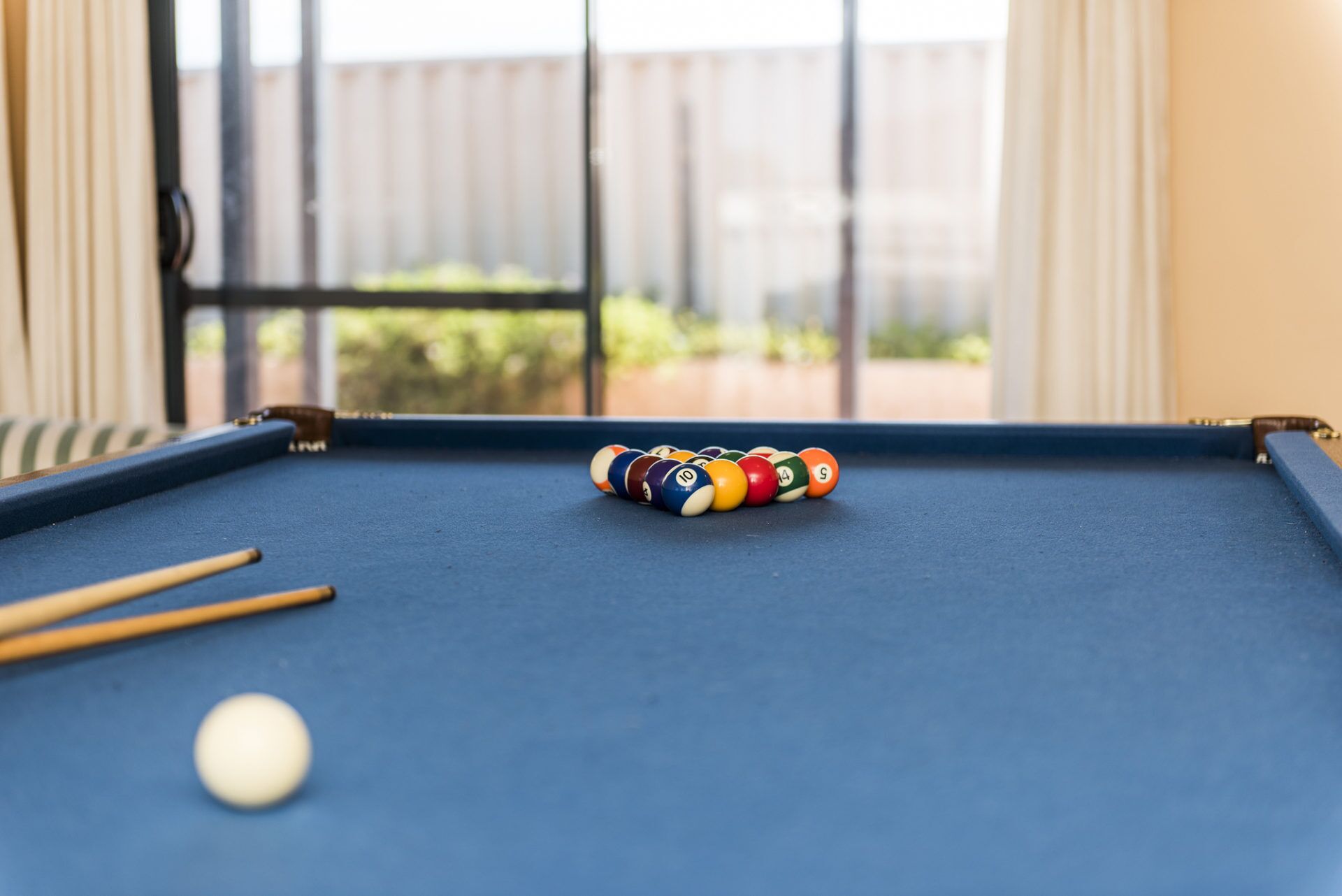 A pool table with a set of billiard balls and cues, a white cue ball, and a window with a view of greenery.