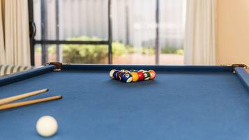 A pool table with a set of billiard balls and cues, a white cue ball, and a window with a view of greenery.