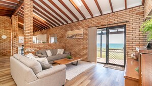 A spacious living room with a brick wall, wooden beams, a sofa, a coffee table, and a sliding glass door leading to a balcony with a view. - Ocean View Retreat - Best Views in town (Guilderton)