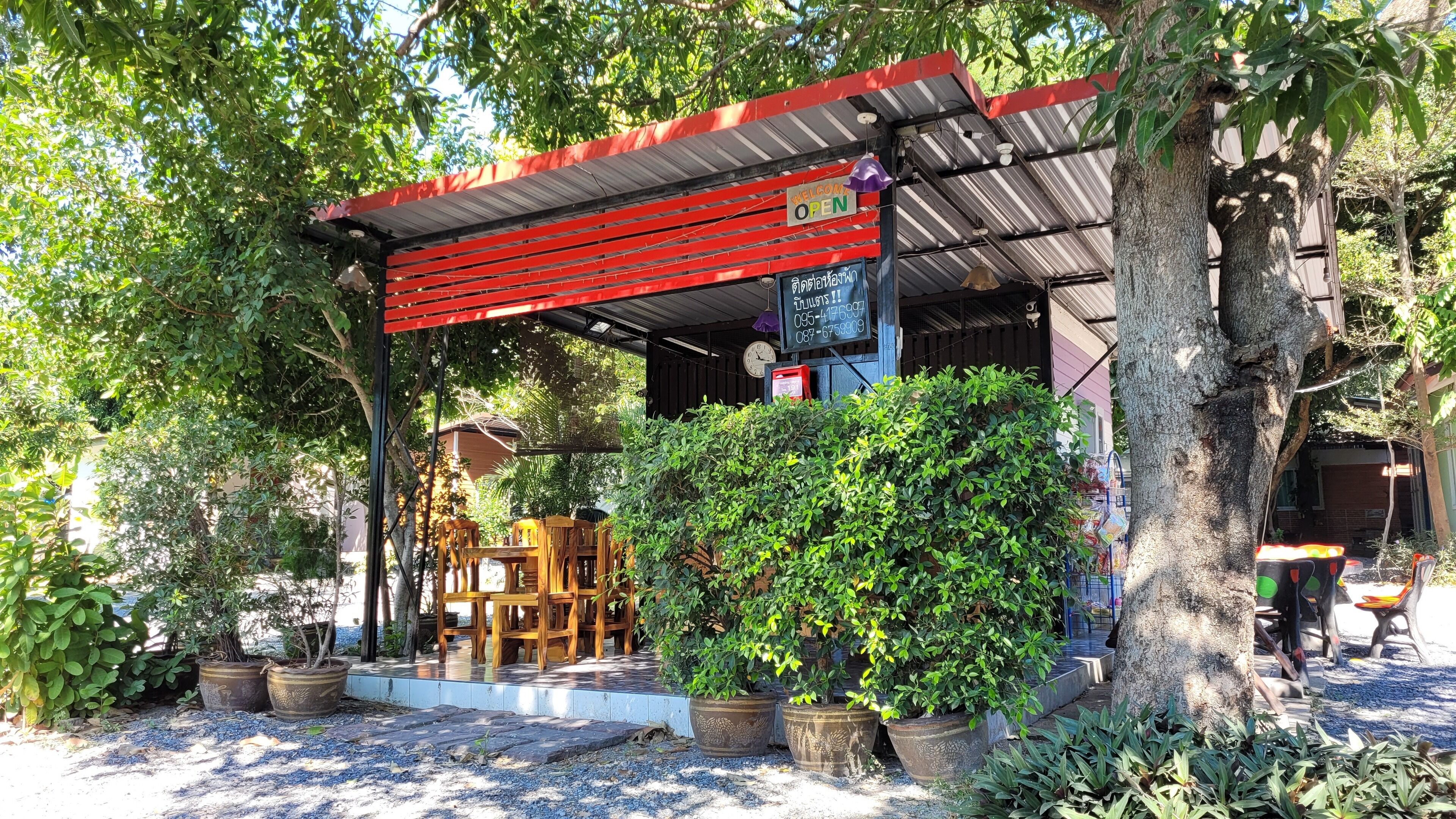 A covered outdoor seating area with wooden furniture, surrounded by potted plants and trees.
