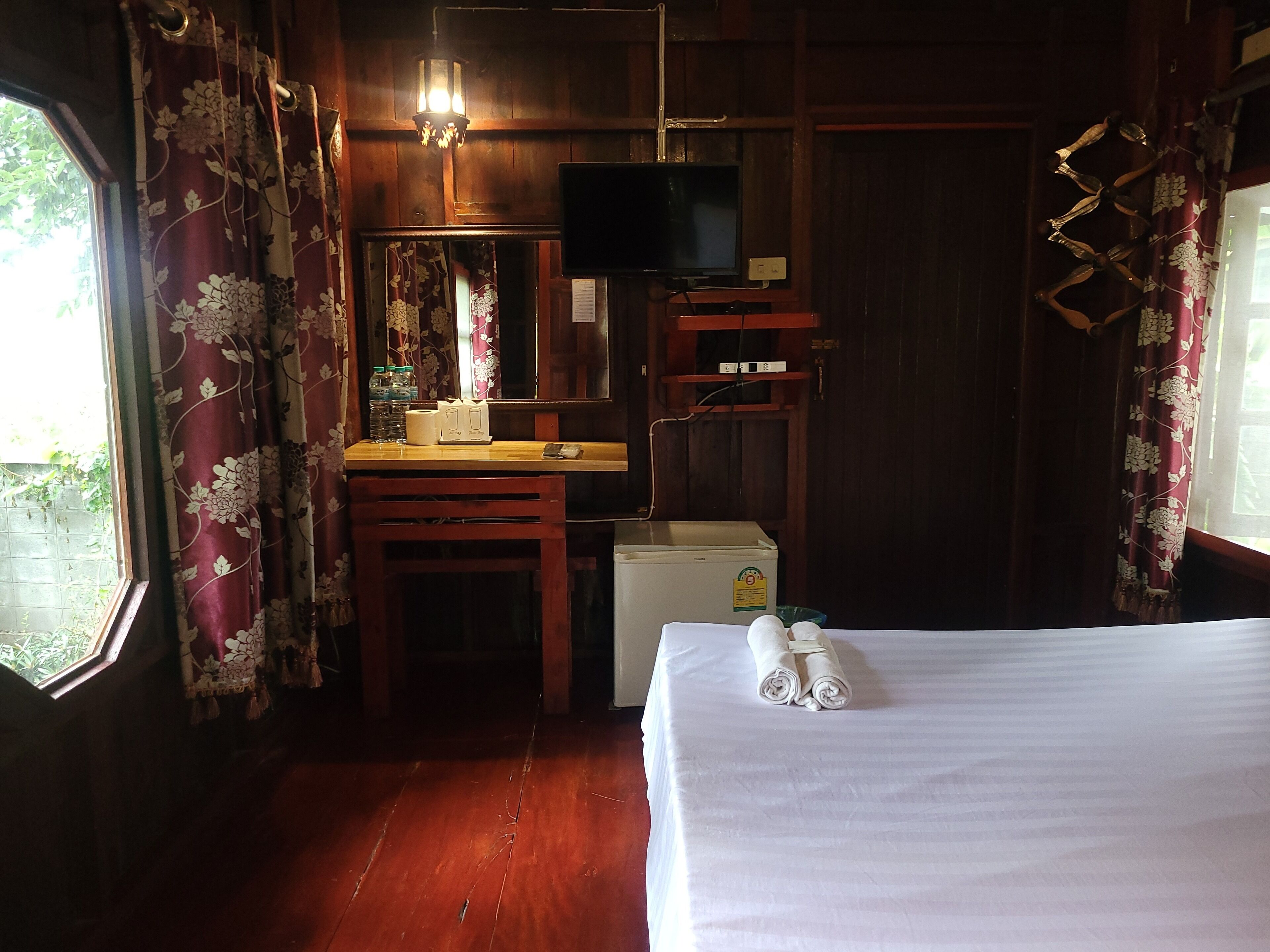 A room with a wooden desk, a television, a bed with towels, and a window with curtains.