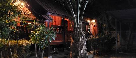 A traditional wooden house with a thatched roof, surrounded by lush greenery and a gravel pathway.