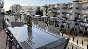Een balkon met een glazen tafel en stoelen, een bloempot en uitzicht op een straat met gebouwen en palmbomen.