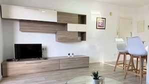 Een moderne woonkamer met een flatscreen televisie, houten opbergmeubels, een glazen salontafel en witte stoelen.