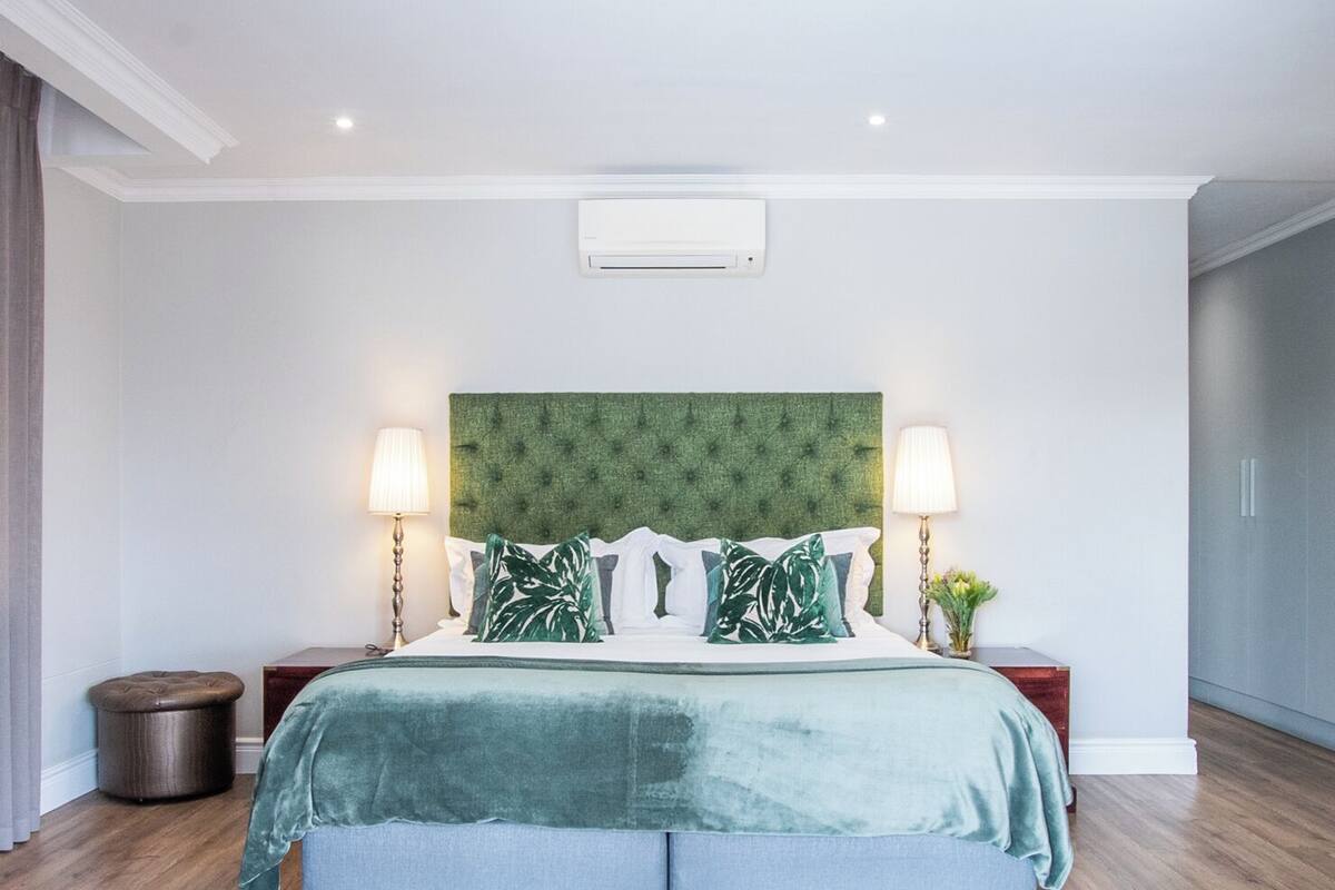 Une chambre moderne avec une tête de lit verte en tissu, un grand lit recouvert d’une couverture verte et blanche, deux lampes de chevet et un grand tapis coloré.
