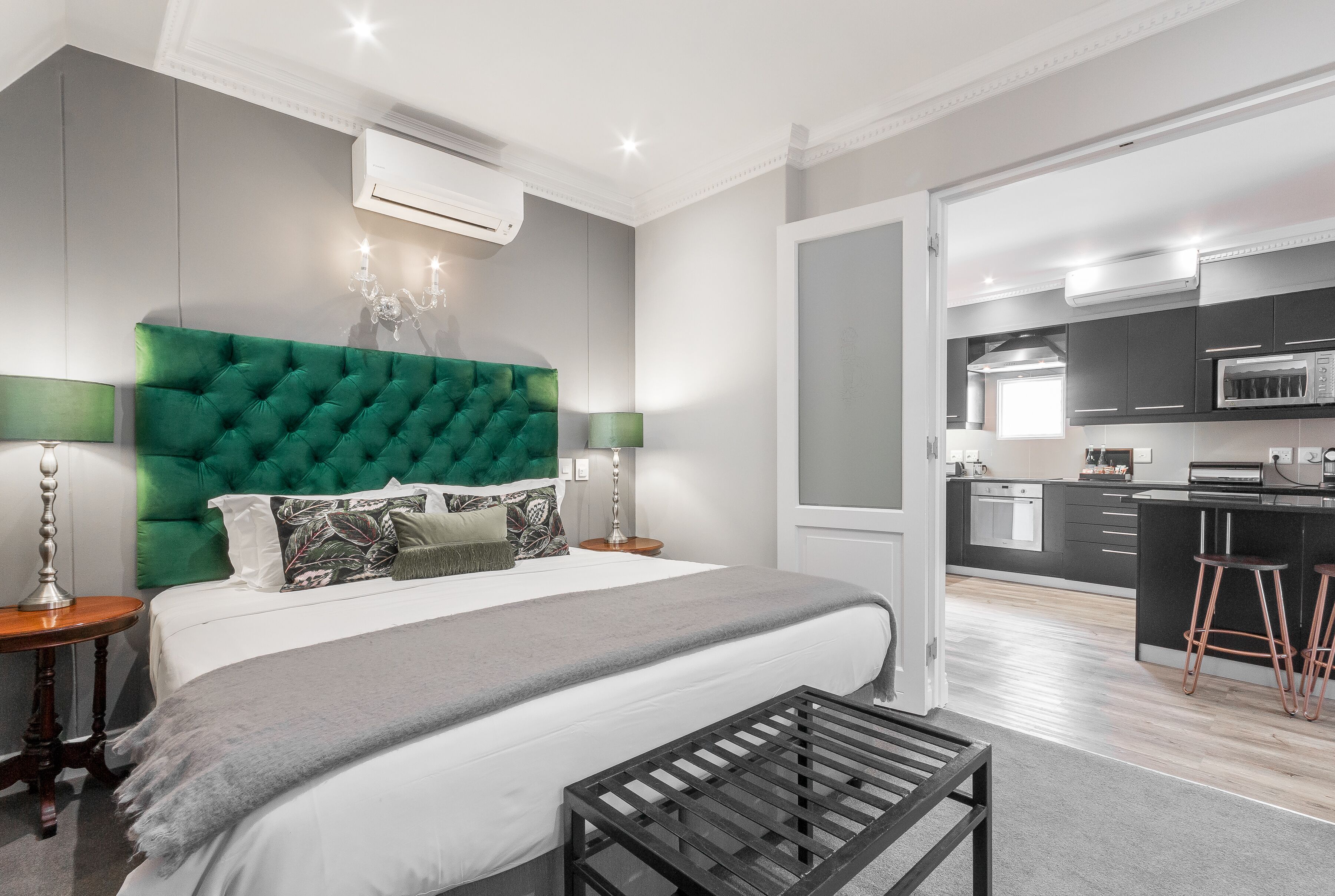 Quarto moderno com cama grande, cabeceira estofada verde, abajures e vista para uma cozinha com um banquinho.