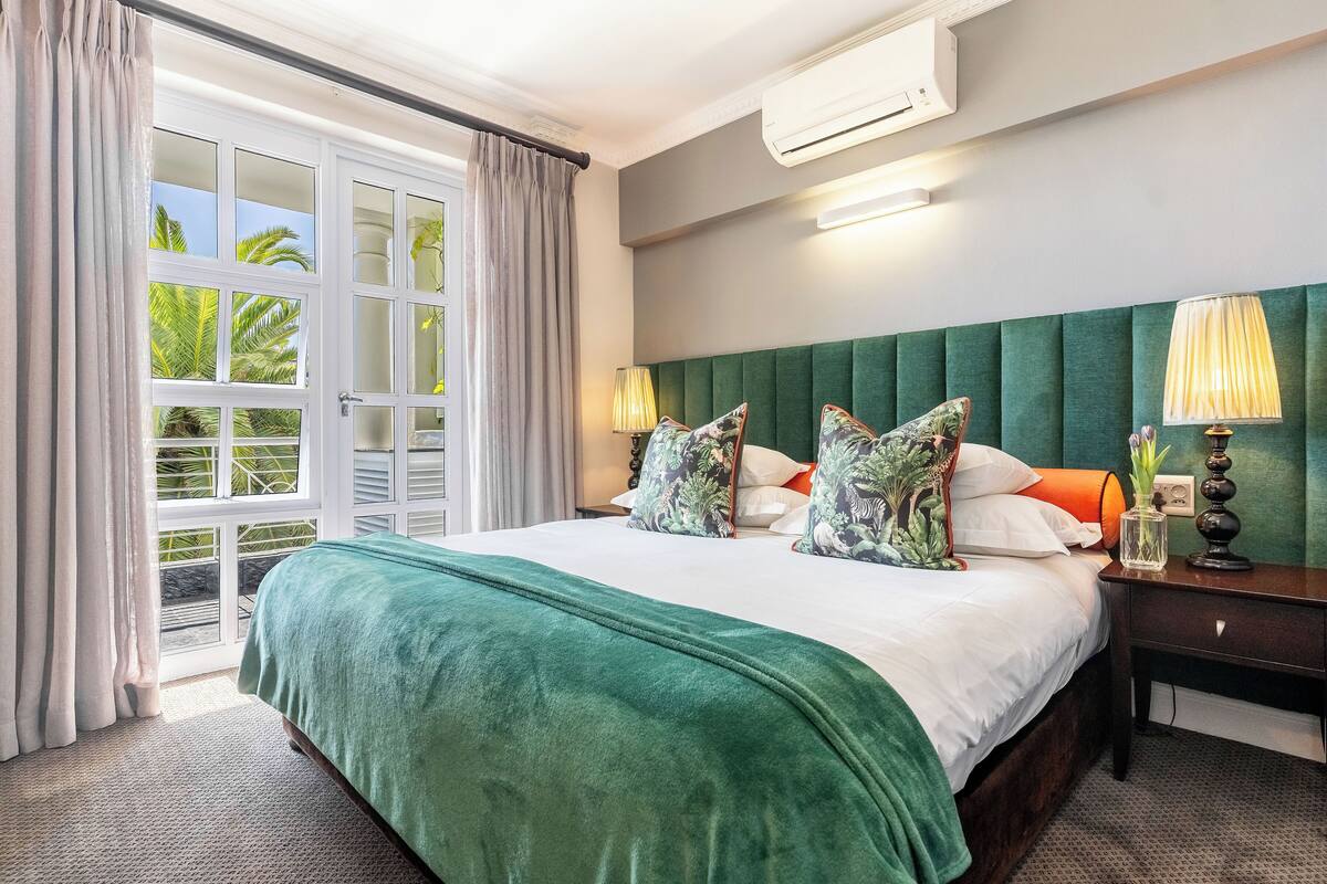Une chambre d’hôtel avec un grand lit, une tête de lit verte, deux lampes de chevet et une vue sur les palmiers, visible à travers une porte-fenêtre coulissante.