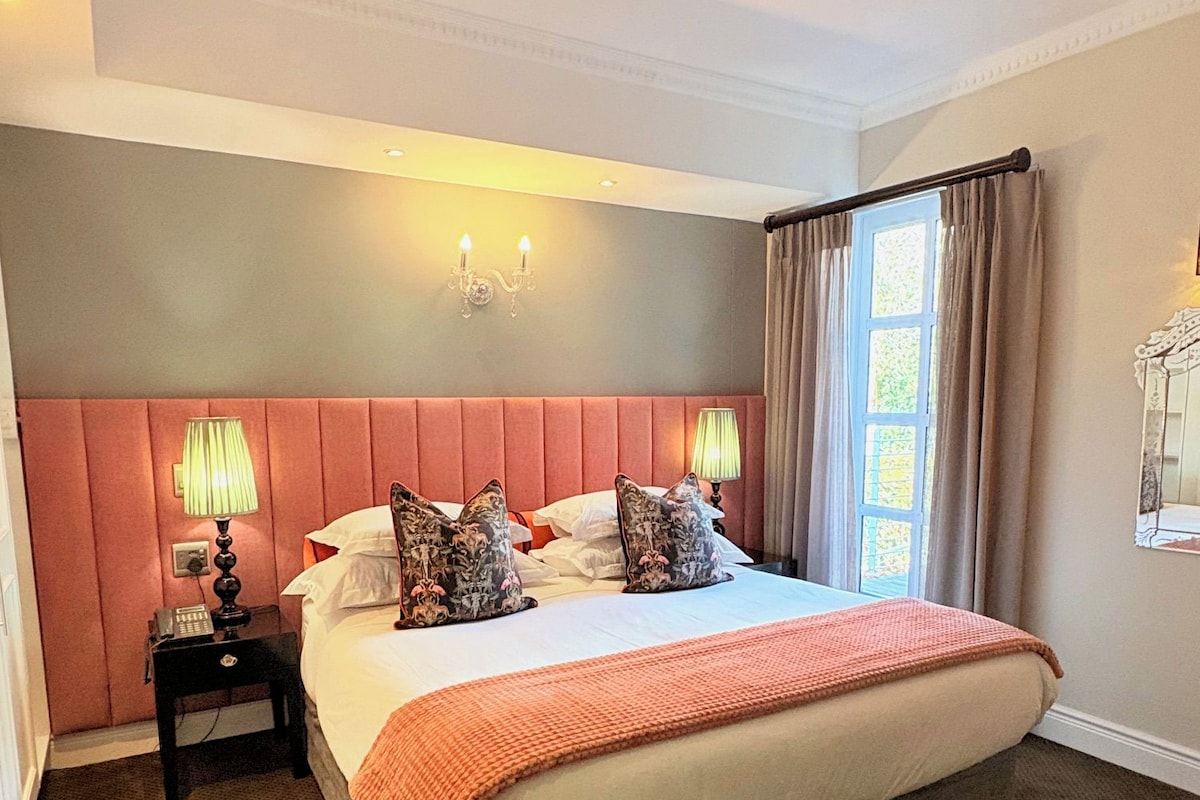 A hotel room with a large bed, a nightstand, a lamp, a wall-mounted light fixture, and a window with curtains.