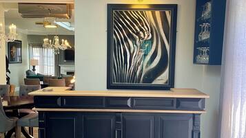 Un salon moderne avec un bar en bois foncé, une grande peinture abstraite et un coin repas avec des chaises en osier.