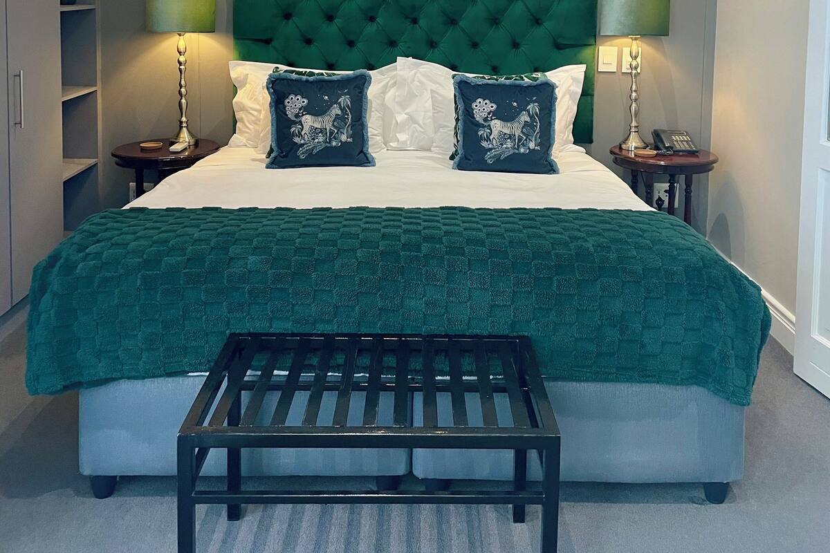 Une chambre à coucher bien aménagée, avec un grand lit, une tête de lit recouverte d’un tissu vert, deux lampes de chevet, une table de chevet avec un téléphone et un petit banc placé devant le lit.