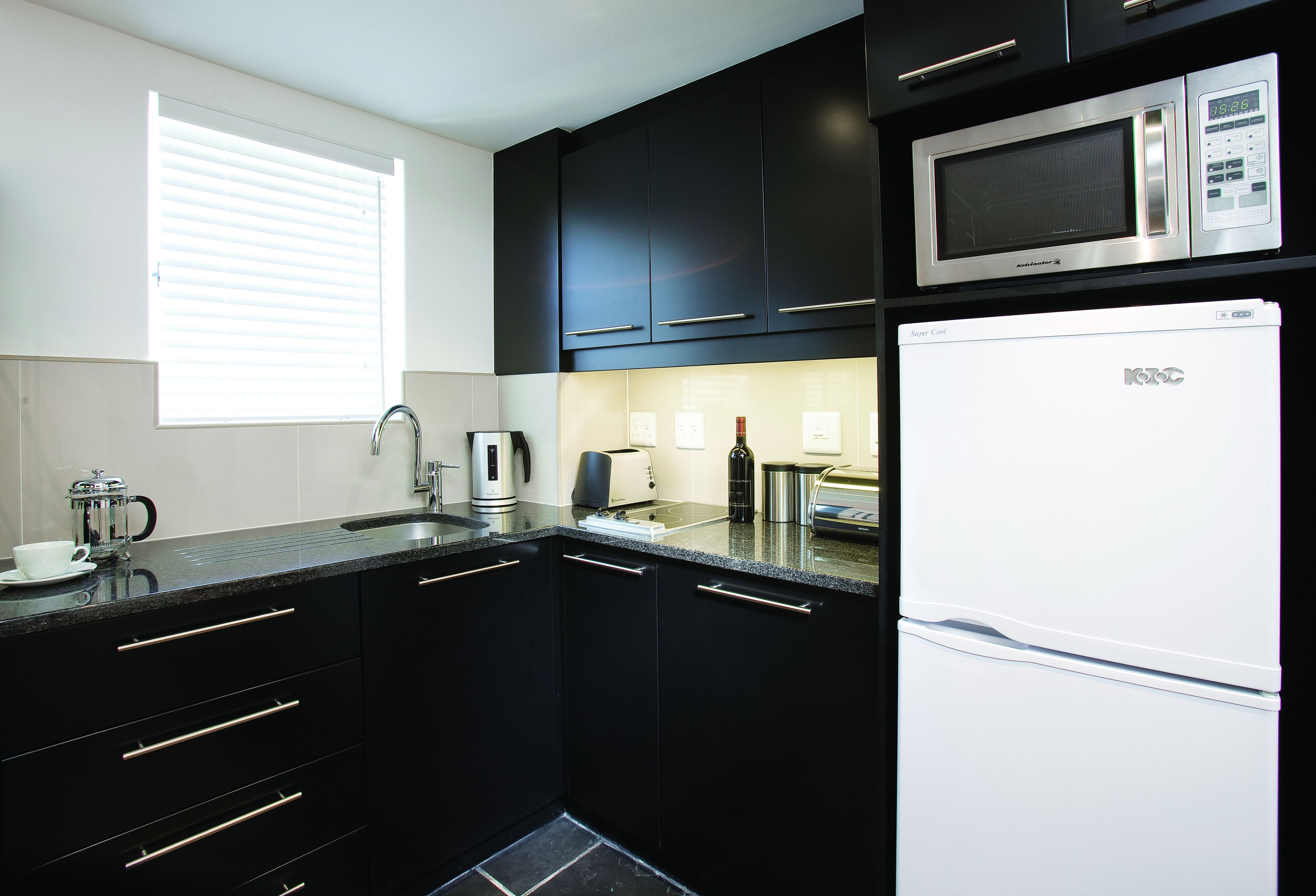 A modern kitchen with black cabinets, a stainless steel microwave, a white refrigerator, and a window with blinds.