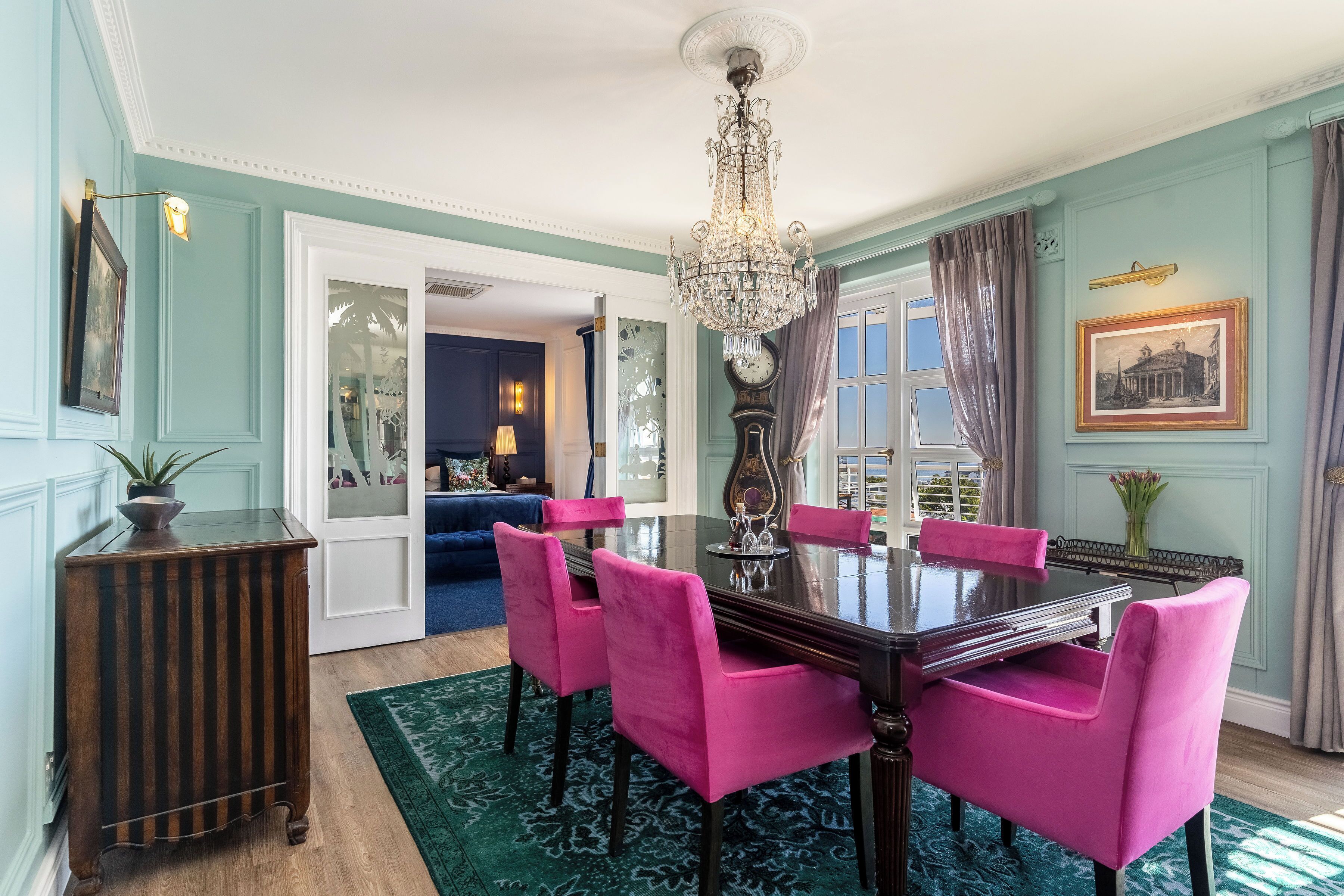 A dining room with a dark wooden table and pink chairs, a chandelier, and a clock on the wall.