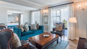 Un salon spacieux avec un canapé bleu, une table basse en bois et un fauteuil noir. On aperçoit un lit avec une tête de lit blanche au fond.