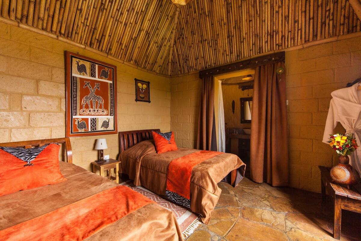 Una habitación con dos camas, cabecera de madera, pared de ladrillo, techo de bambú y una puerta que da a otra habitación.