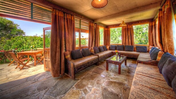 A cozy interior with a stone floor, wooden furniture, and large windows offering a view of greenery. - Rhino Watch safari Lodge (Nyahururu)