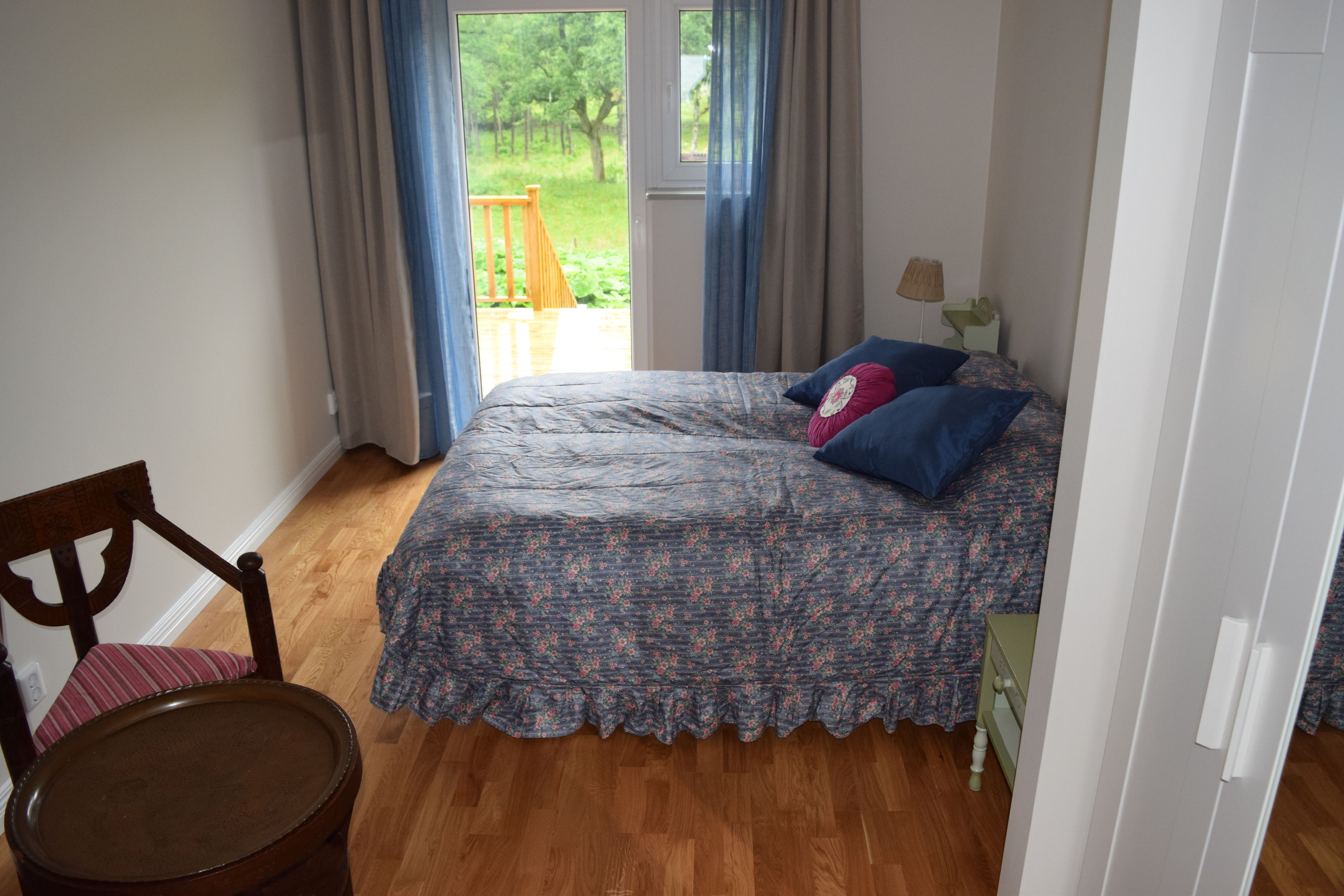 A bedroom with a wooden floor, a bed with a floral bedspread, a chair, and a window with curtains.