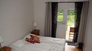 Une chambre Ă coucher avec un lit, une table de chevet, une lampe et une vue sur un jardin, visible Ă travers une porte.