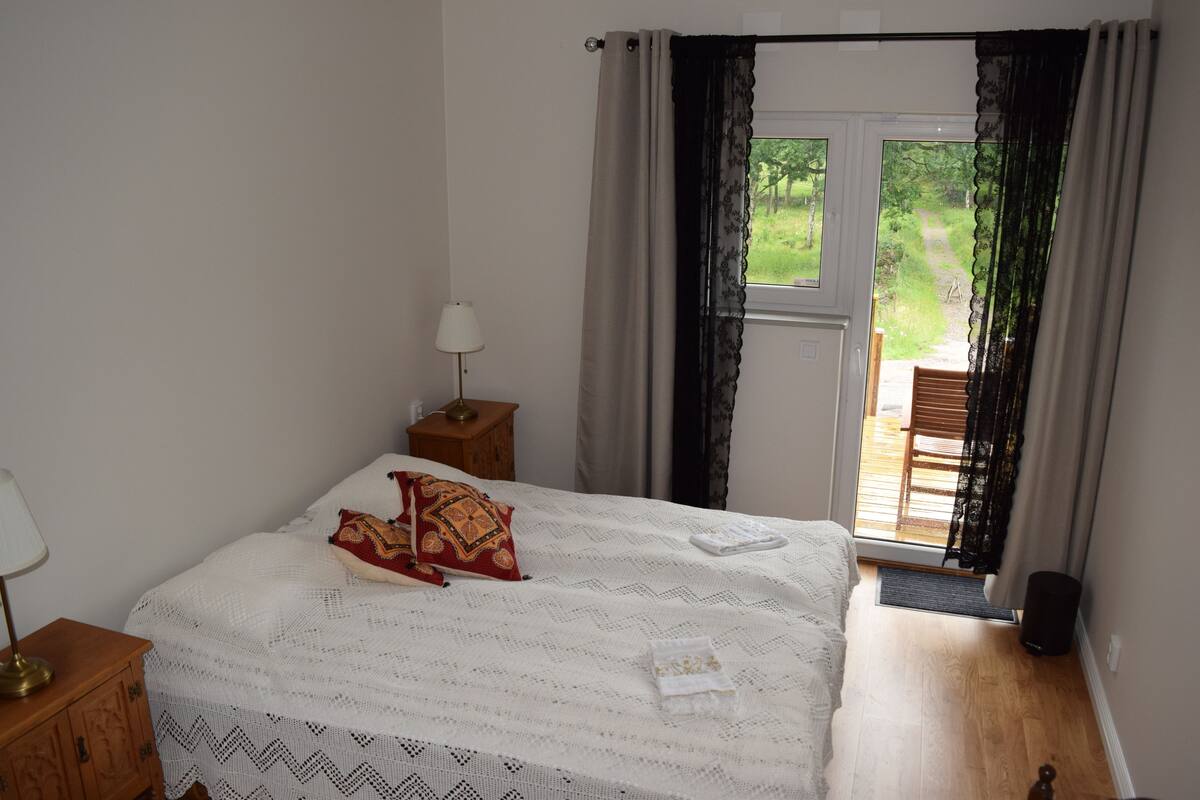 A bedroom with a bed, a nightstand, a lamp, and a view of a garden through a door.