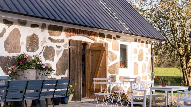 Un bâtiment en pierre avec une porte en bois, un espace extérieur abrité avec des bancs et des tables, et des plantes en pot.