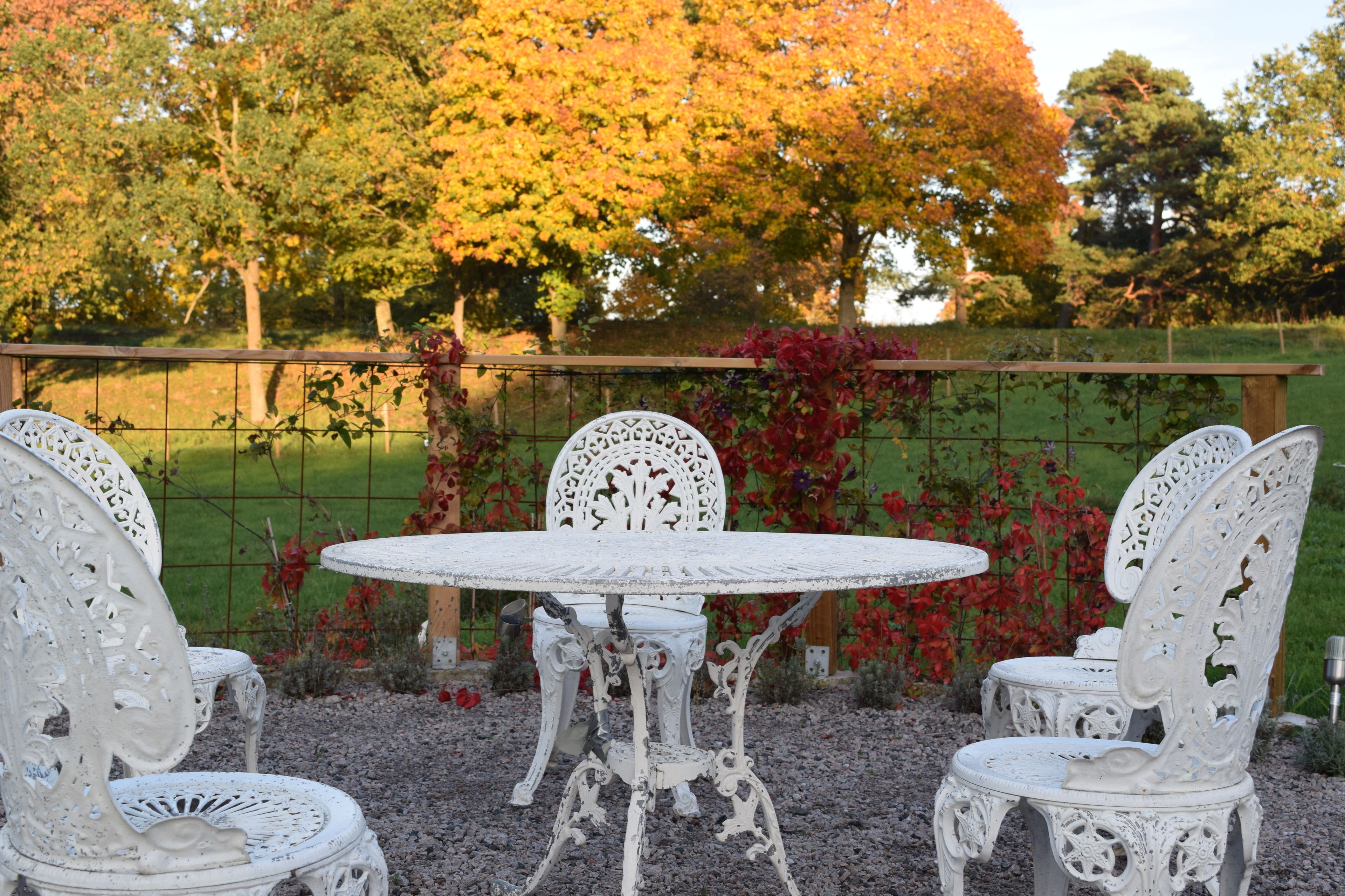 A white outdoor dining set with a round table and four chairs, surrounded by a gravel area and a fence with greenery and trees in the background.