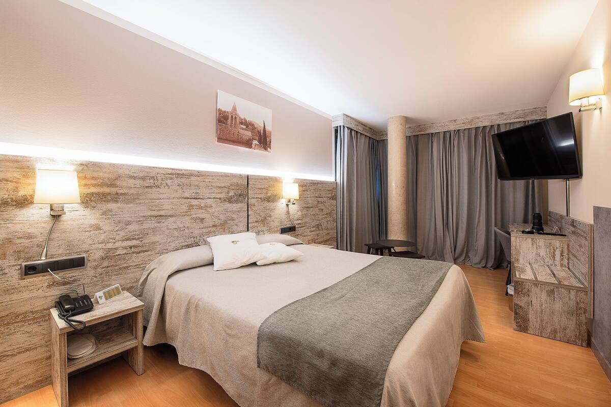 Une chambre d’hôtel moderne dotée d’un grand lit, d’une télévision à écran plat, d’une table de chevet avec une lampe et d’une prise de courant.
